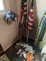Photo showing multiple American flags on metal and wooden poles standing in corner, some flagged are folded or draped, with packaged garden flags on floor.