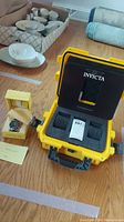Yellow Invicta watch case, Invicta watch in box, extra band links, and paperwork on wood floor