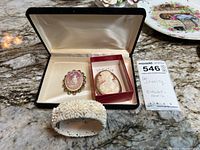 Photo showing two cameo brooches and a carved shell bracelet placed on a marble surface with a jewelry case and auction tag.