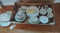 Box with organized assortment of dinnerware pieces including plates, cup and saucer sets, bowls, small trays, and bowls with floral pattern and silver trim.