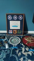 Wide shot showing the framed certificate with NRA instructor patches along with several belt buckles, a plate, and a plaque arranged on the carpeted floor.