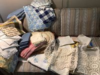 Overall photo displaying stacks of folded crocheted and cut work linens, napkins, and towels on a couch showing condition and variety.