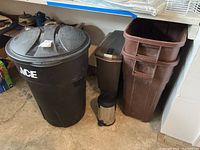 Three Rubbermaid Rough Neck outdoor trash cans with lids, two indoor foot pedal trash cans (one black rectangular, one stainless steel cylindrical) grouped together in garage floor area.