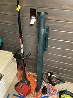 Craftsman gas weed trimmer, electric hedge trimmer, BD cordless broom, and extension cord shown together on floor and leaning against wood wall.