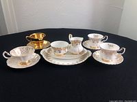 Full lot with 3 tea cups and saucers, one gold, one white with gold 50th anniversary detail, creamer, and sugar dish on black background.