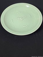 Full view of the round celadon glazed plate showing the light green matte surface with raised dragon and fish reliefs along with swirling pattern decorations.
