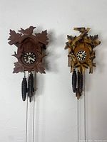 Front view of two wooden carved cuckoo clocks showing bird and leaf motifs, pinecone weights, and chains.