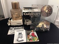 Photo showing the assembled Cuisinart DLC-7 food processor with power cord, pasta maker box, acrylic accessory holder containing three pasta discs, and three cookbooks/manuals arranged on black table.