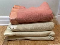 Three wool blankets stacked: pink top blanket with satin pink trim, middle cream colored blanket with ivory satin trim, bottom larger cream blanket with satin trim.