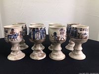Seven off-white stoneware chalices with painted Egyptian motifs standing in a row on a black surface, shown from the front and slightly above to capture details on the bowl part and stemware shape.