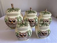 Front view of five vintage owl ceramic canisters with lids showing detailed owl faces and leafy vine decorated lids topped with owl figurine handles.