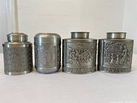 Front view of four pewter lidded canisters showing embossed Asian motifs including calligraphy and figures
