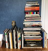 Stack of approximately 40 photography and related books on a shelf against a blue wall and white surface, showing titles including The Story of Kodak and The Darkroom Book.