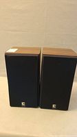 Pair of speakers with black fabric front covers and wood veneer sides, standing upright.