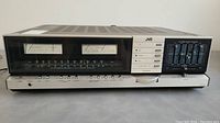 Front view of JVC JR-S401 stereo receiver showing large VU meters, analog FM/AM tuner display, source selector buttons, and 10-band graphic equalizer with sliding volume control.