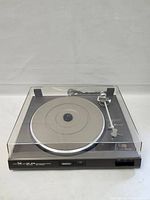 Side angled top-down photo of the DUAL CS 515 turntable inside its clear dust cover showing platter, tonearm with Ortofon cartridge, and control buttons on the front.