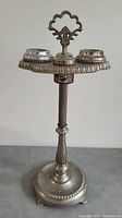 Front view of vintage metal standup ashtray showing handle, three ash cups and decorative top rim.