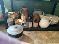 Shelf display showing two white scalloped bowls, two small stoneware crocks, two pitchers and decorative vase