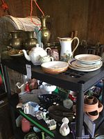 Overall view of two-shelf black plastic cart holding assortment of brass, porcelain, glass items including vases, plates, teapot, pitchers, and small figurines.