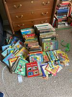 Photo showing assorted children's books spread on floor including Little Golden Books and other classical titles near a wooden chest of drawers.