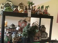 Photo showing top shelf with nativity figures, palm trees, ceramic houses with snow, and artificial trees on lower shelf