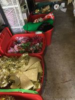 Four large red storage totes filled with assorted Christmas decorations including gold glitter bows, artificial flowers, and garlands.