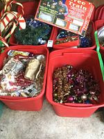 Four large red totes containing various Christmas decorations including glass ornaments, garland, candy canes, and a boxed tree train set.
