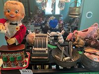 Wide shot showing vintage electronic bartender toy in a red jacket with a shaker and glass, mechanical metal calculator, white and brown rabbit figurines, and a black bowl holding razors. Also visible are various other collectibles in the background.