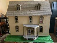 Front exterior view of the large doll house showing detailed textured walls, arched windows, front porch with double doors, and shingled roof with two dormers. The house is on a green base resembling grass.
