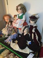 Full group of various dolls including one large doll with green dress and two dolls in burgundy velvet dresses with lace, placed on a shelf.