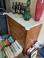 Front and side view of the wood dresser with stone top, showing brass handles, faux keyholes, and surrounding area with books and decor items. Dresser is polished medium wood tone with detailed legs.