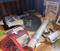 Photo showing a pile of assorted historical newspapers, magazines, papers, rolled maps or posters, and a large oval vintage photograph of a seated man.