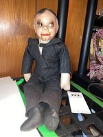 View of vintage ventriloquist dummy sitting against wooden background, showing full body from front with facial details and suit.