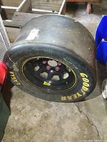 View of flat Goodyear Eagle race tire mounted on a black steel rim with yellow striping and pink lug nuts, tire surface smooth and worn