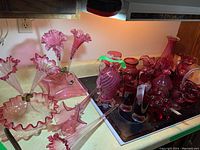 Wide view of multiple red and cranberry glass pieces arranged on a kitchen counter, including vases, bowls, epergnes, glasses, and decorative glass items.