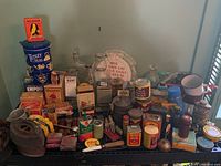 Wide view of the lot showing sad irons, blue Tetley Teas tin, various vintage boxes, enamel cups, wooden pegs, and glass bottles