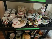 Eight floral mugs, five tea pots including a decorative bunny shaped piece, white plates and cups stacked, and a colorful pitcher.