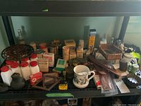 Overview photo showing multiple milk glass bottles with red metal lids, various vintage first aid and baby powder containers, a mustache cup, metal lids and kitchen accessories on a black plastic shelf.
