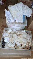 Top view of two plastic storage bins filled with vintage textiles, including a white blanket with pastel embroidery and lace doilies, gloves and fabric pieces.