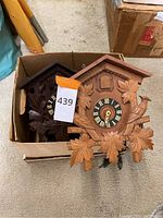 Photo of two cuckoo clocks in a box showing traditional carved wooden design with clock faces and detached parts.