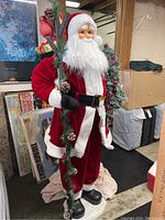 Front view of the Santa Claus figure wearing red and white suit, holding decorated evergreen branch and small Christmas tree.