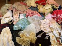 Photo showing multiple baby clothes including dresses with lace trims, pants, and crocheted items in varying pastel colors and floral prints