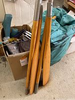 Wooden oar shafts and blades leaning against a teal bag and cardboard box