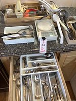 Overview of stainless steel flatware including forks, spoons, knives arranged in white plastic trays, metal ladles, plastic serving utensils, and kitchen tools on countertop