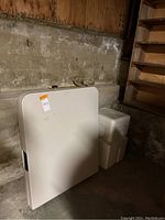 White folding table leaning against a concrete wall in a garage, with a white cooler beside it.