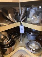 Kitchen corner cabinet with stainless steel lids on top shelf, Cuisinart processor parts including blades and containers on right