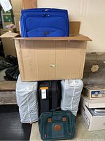 Photo showing the lot of 4 suitcases and 1 bag stacked with two white-covered items, a blue suitcase, black suitcase, and green bag visible.