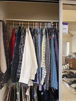 Closet with men's shirts hung on hangers in various colors and patterns, ties hung on side, fleece jackets hung below, and women's pants and packaged clothing on floor.