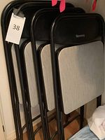 Four black Samsonite folding chairs with beige Grace fabric seats, compact size, folded and stacked against wall.