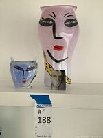 Three glass decorative pieces displayed on a shelf: blue cat face sculpture, pink cat face vase, and small rectangular face sculpture